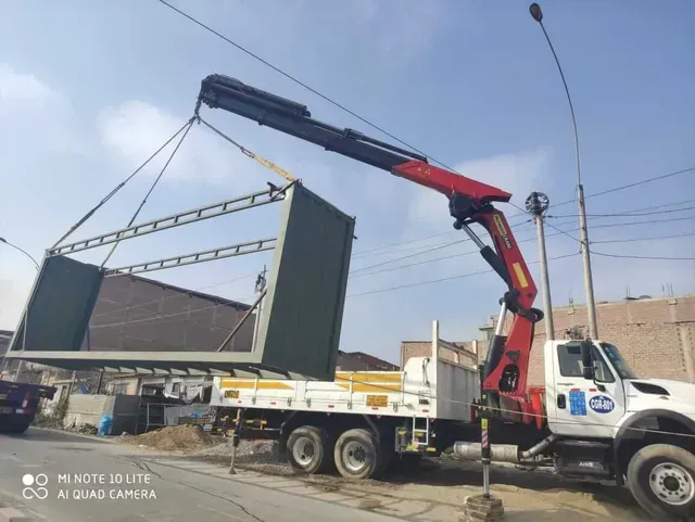 Una grúa montada sobre un camión levanta un gran panel de metal oscuro en una obra de construcción bajo un cielo despejado.