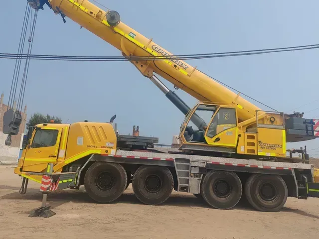 Camión grúa amarillo con pluma extendida estacionado en tierra bajo líneas eléctricas