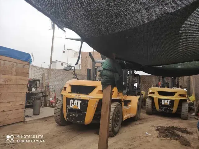 Carretillas elevadoras CAT amarillas bajo un toldo negro en un patio industrial, con un trabajador de pie sobre una de ellas.