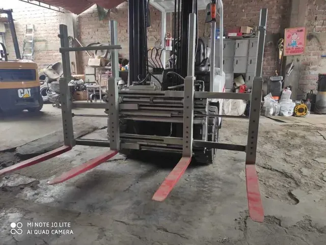 Una carretilla elevadora en el patio de un almacén en mal estado, con largas horquillas rojas extendidas hacia la cámara.