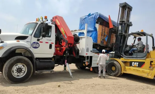 Una grúa y una carretilla elevadora cargan una caja envuelta en papel azul sobre un camión de plataforma en un terreno polvoriento.
