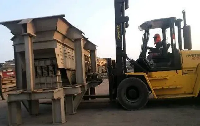 Una carretilla elevadora amarilla mueve una gran tolva metálica industrial en un patio al atardecer.