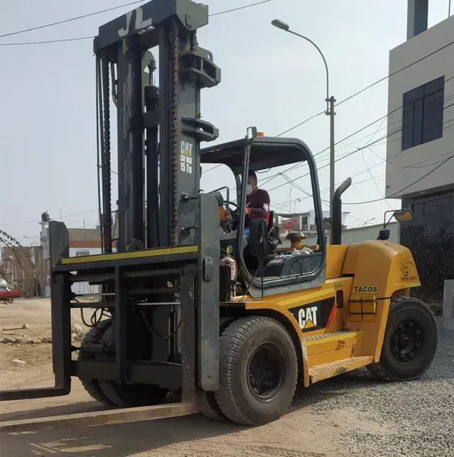 Una carretilla elevadora CAT amarilla levanta un palé en un patio industrial al aire libre con un trabajador en la cabina.