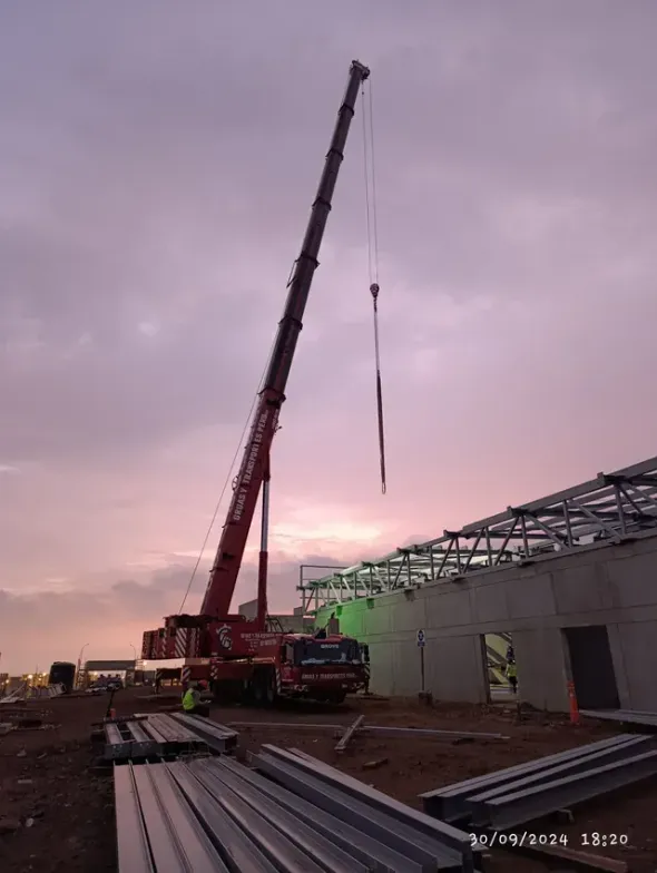 Grúa levantando una viga larga en una obra de construcción al atardecer, con materiales de acero en primer plano.