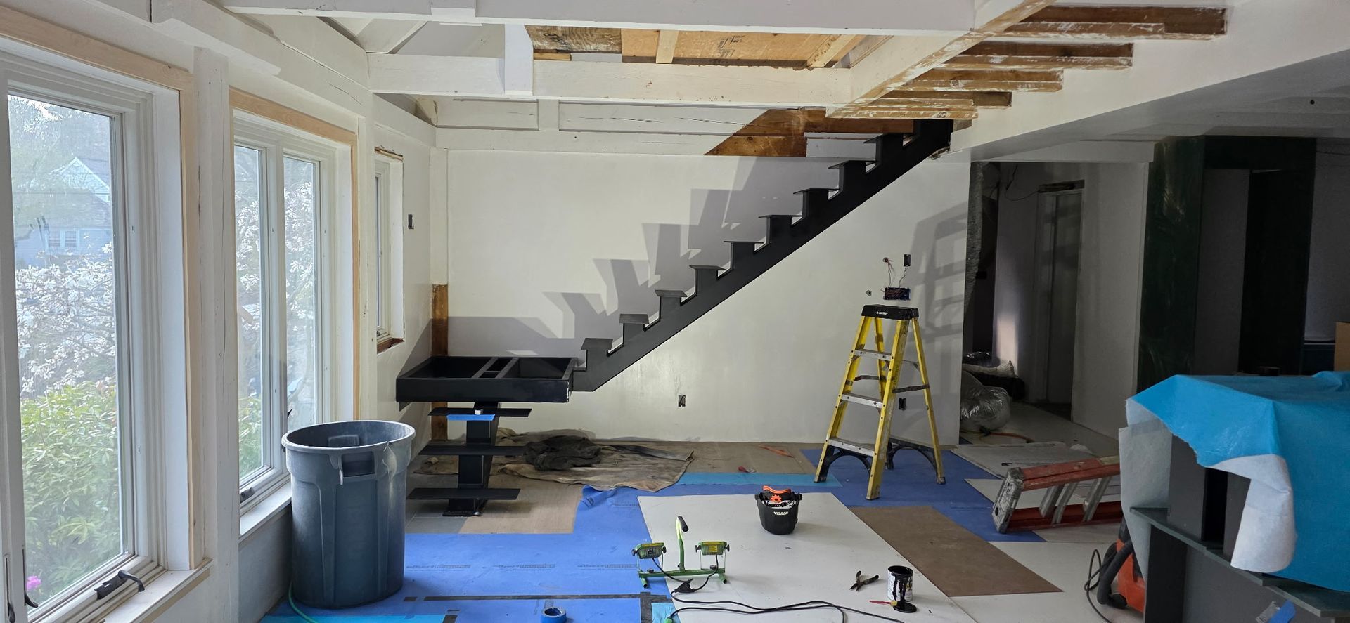 Renovation site with stairs, ladder, blue tarp, and tools in a bright room with large windows