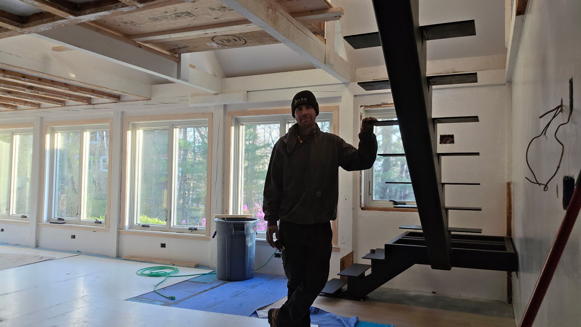Person standing in a bright unfinished room beside a black staircase and large windows.