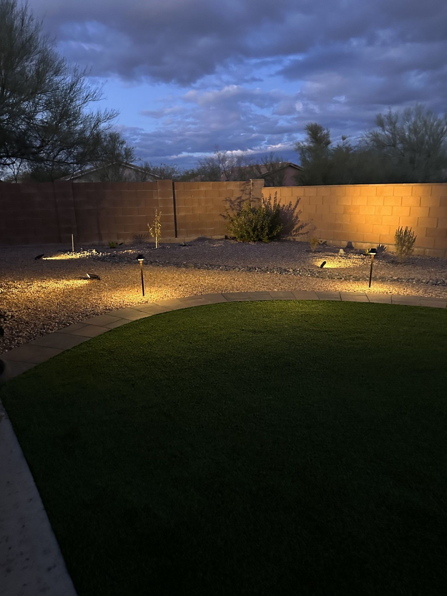 Backyard at dusk with lit landscaping and a lawn; a cloudy sky overhead.
