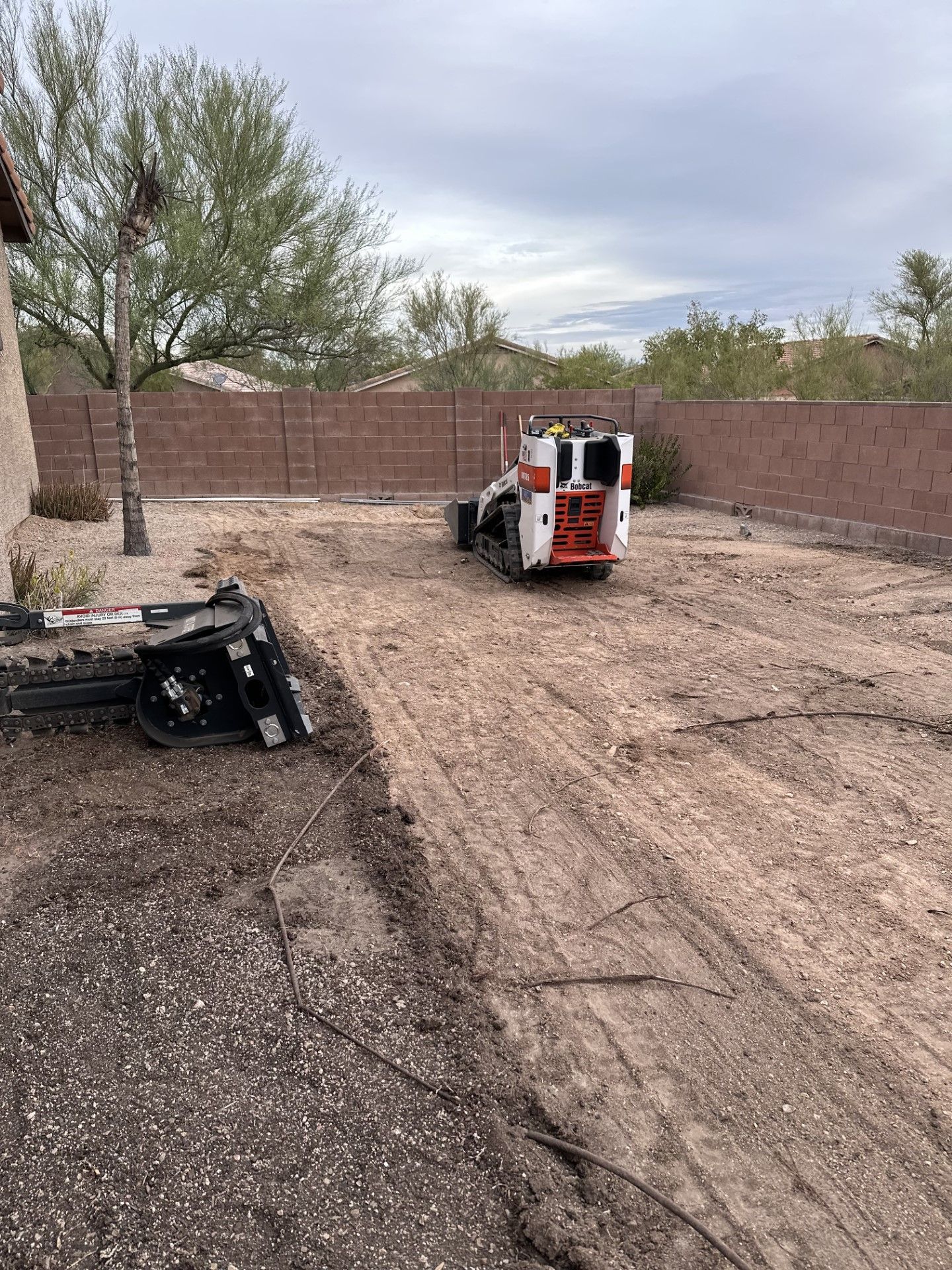 A compact construction vehicle in a dirt backyard clearing. Brown fence and overcast sky.