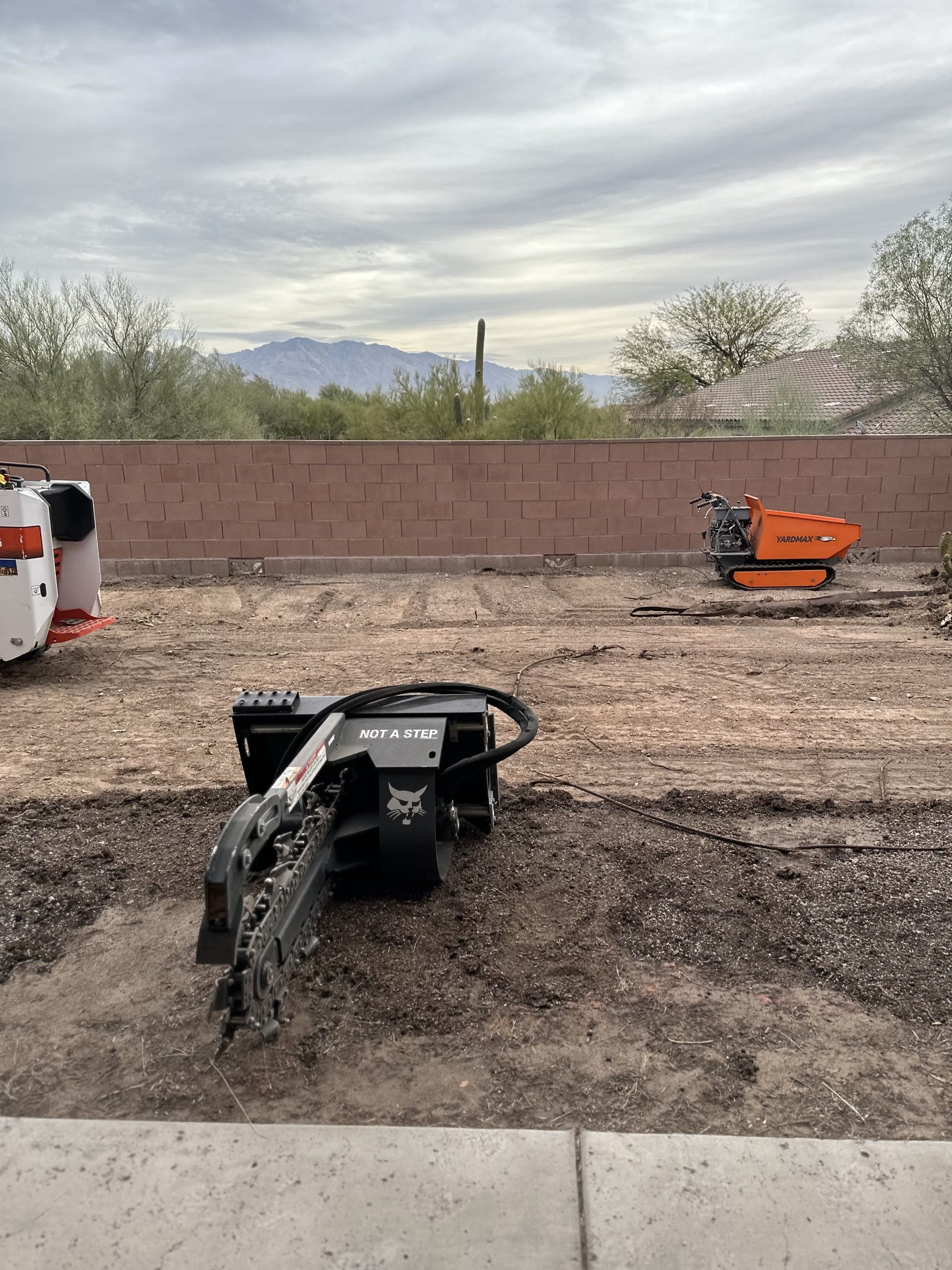 Construction site with three pieces of machinery on dirt, brown wall and mountains in background.