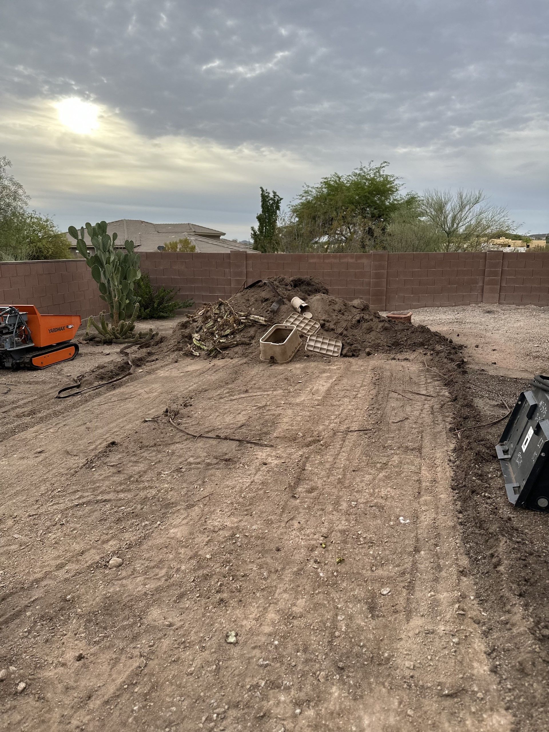 Dirt pile in a yard, equipment present. Brown dirt and gravel ground with a brick wall backdrop under cloudy sky.