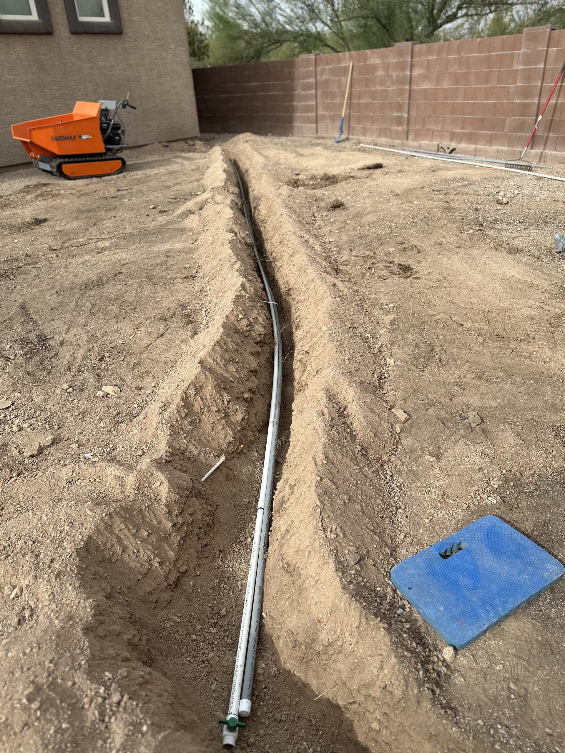 A trench dug in dirt, with buried PVC pipe and a small orange construction vehicle in the background.