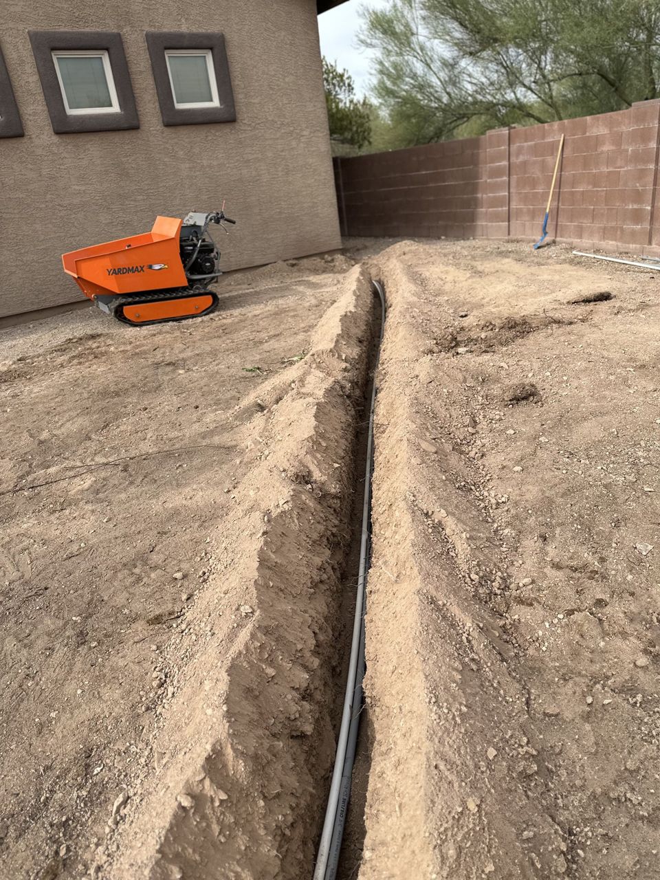 A narrow trench dug in dirt, with a utility vehicle nearby, and a house wall in the background.