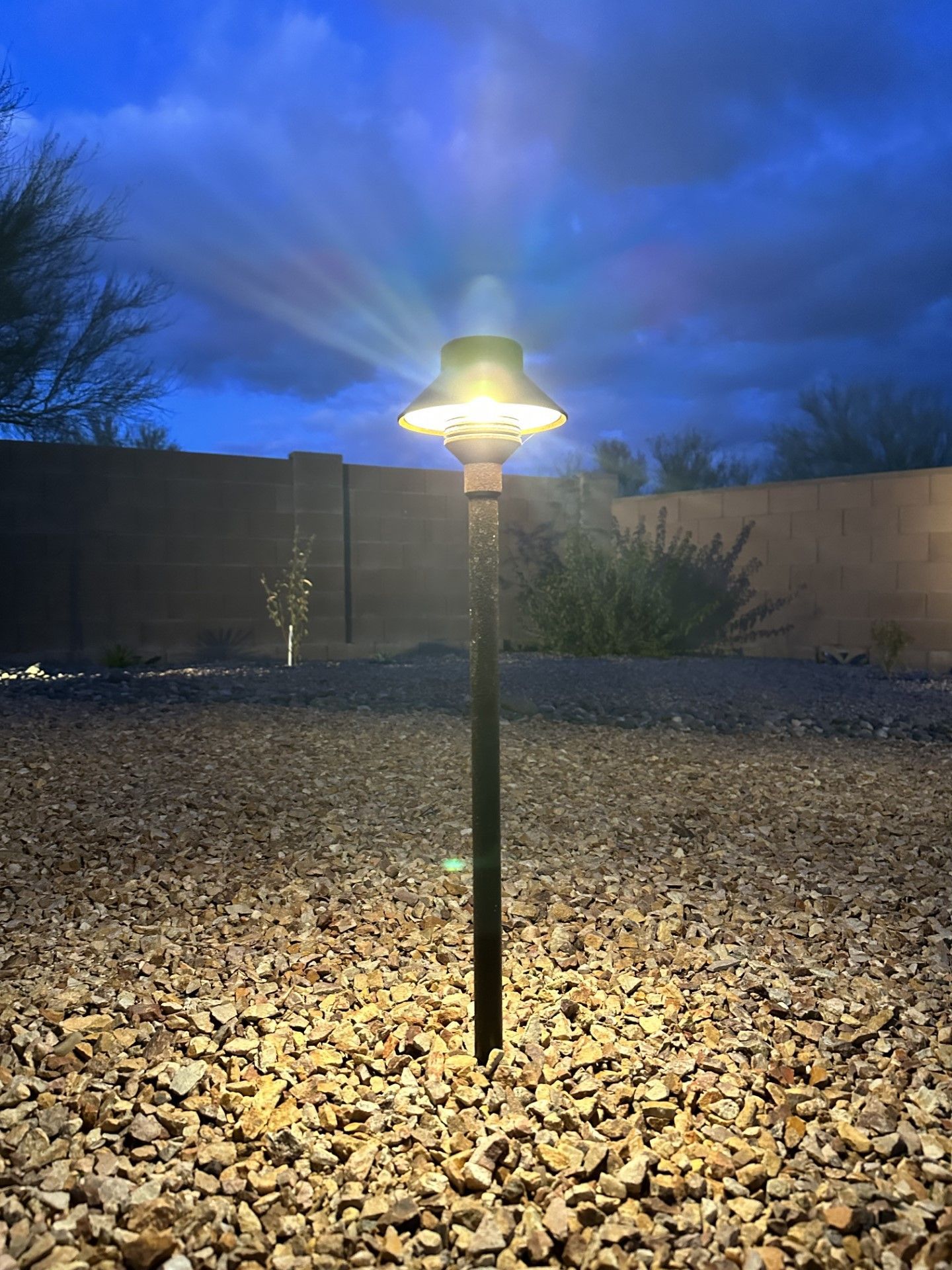 A lit outdoor pathway light against a dusk sky and a gravel yard.