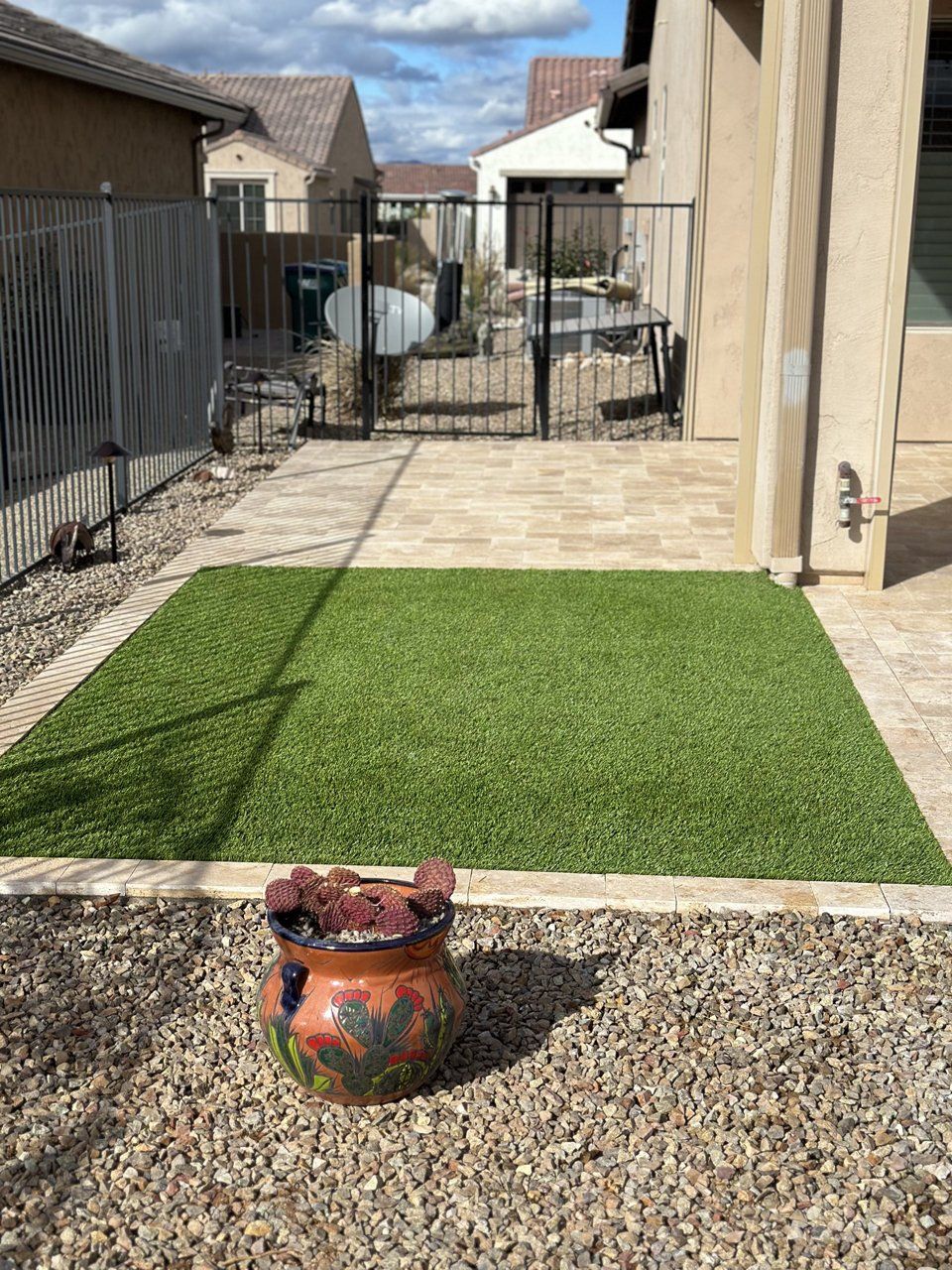 Backyard with artificial turf surrounded by pavers and gravel, a decorative pot, and a black metal gate.