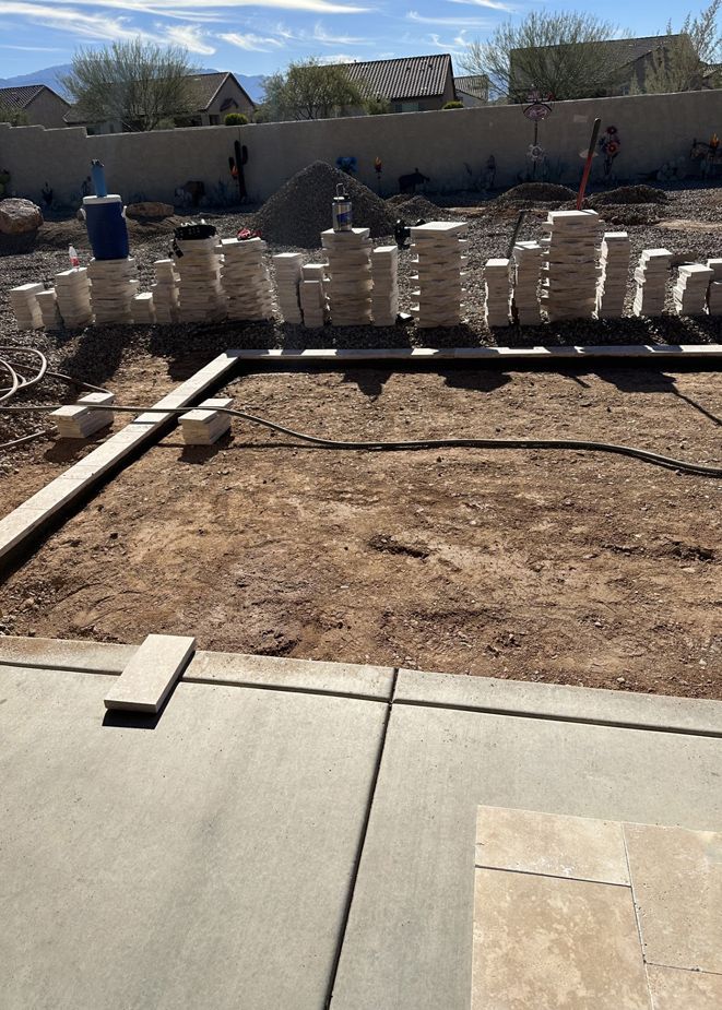 Construction site: Ground marked for patio, surrounded by stacked paving stones, concrete, and a wall.