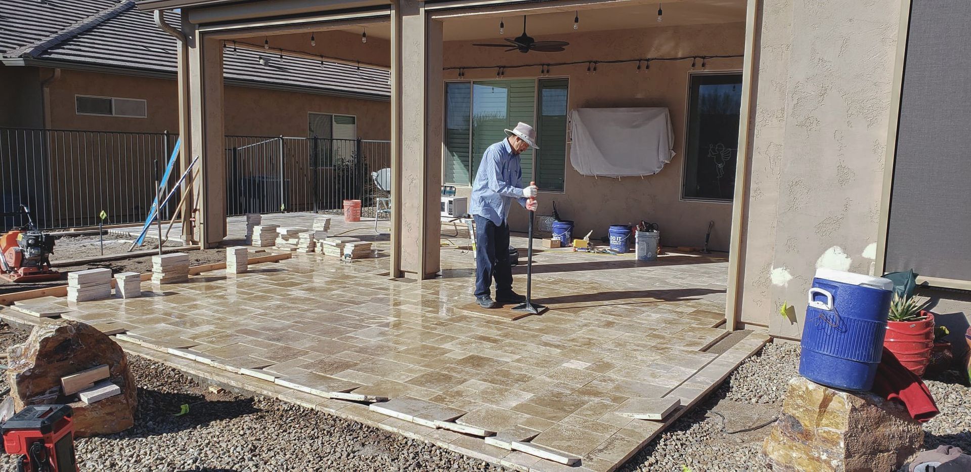 Construction worker leveling wet concrete on a patio. Gray concrete, beige pillars, blue bucket, rocky yard.