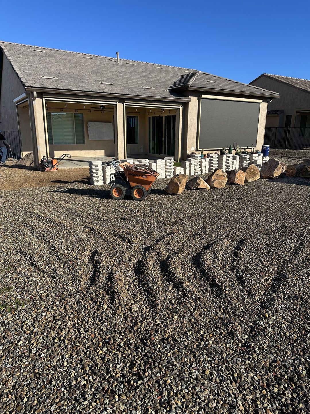 Backyard with gravel ground, small tractor, building with patio cover, and stacked blocks.