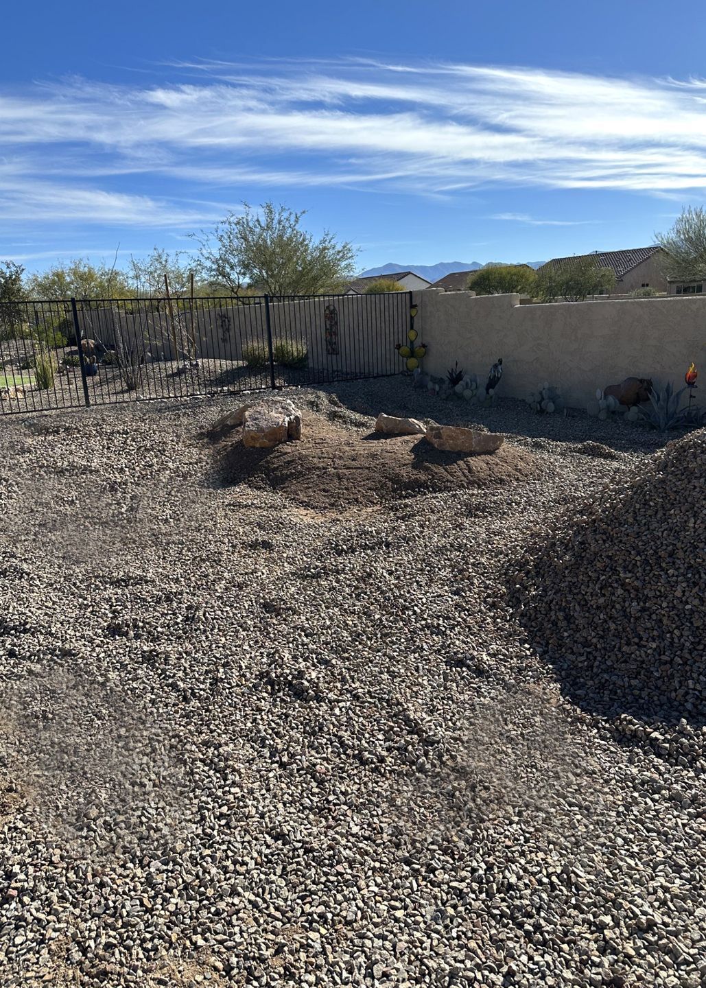 Gravel-covered backyard with a concrete wall and fence under a blue sky with some brush.