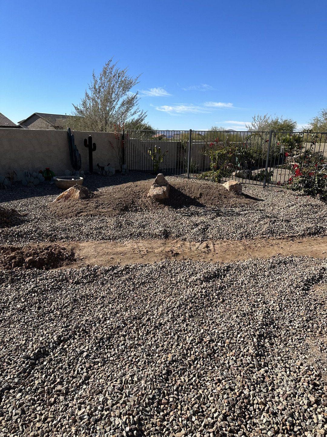 Gravel-covered backyard with dirt mounds and scattered plants against a wall and clear blue sky.