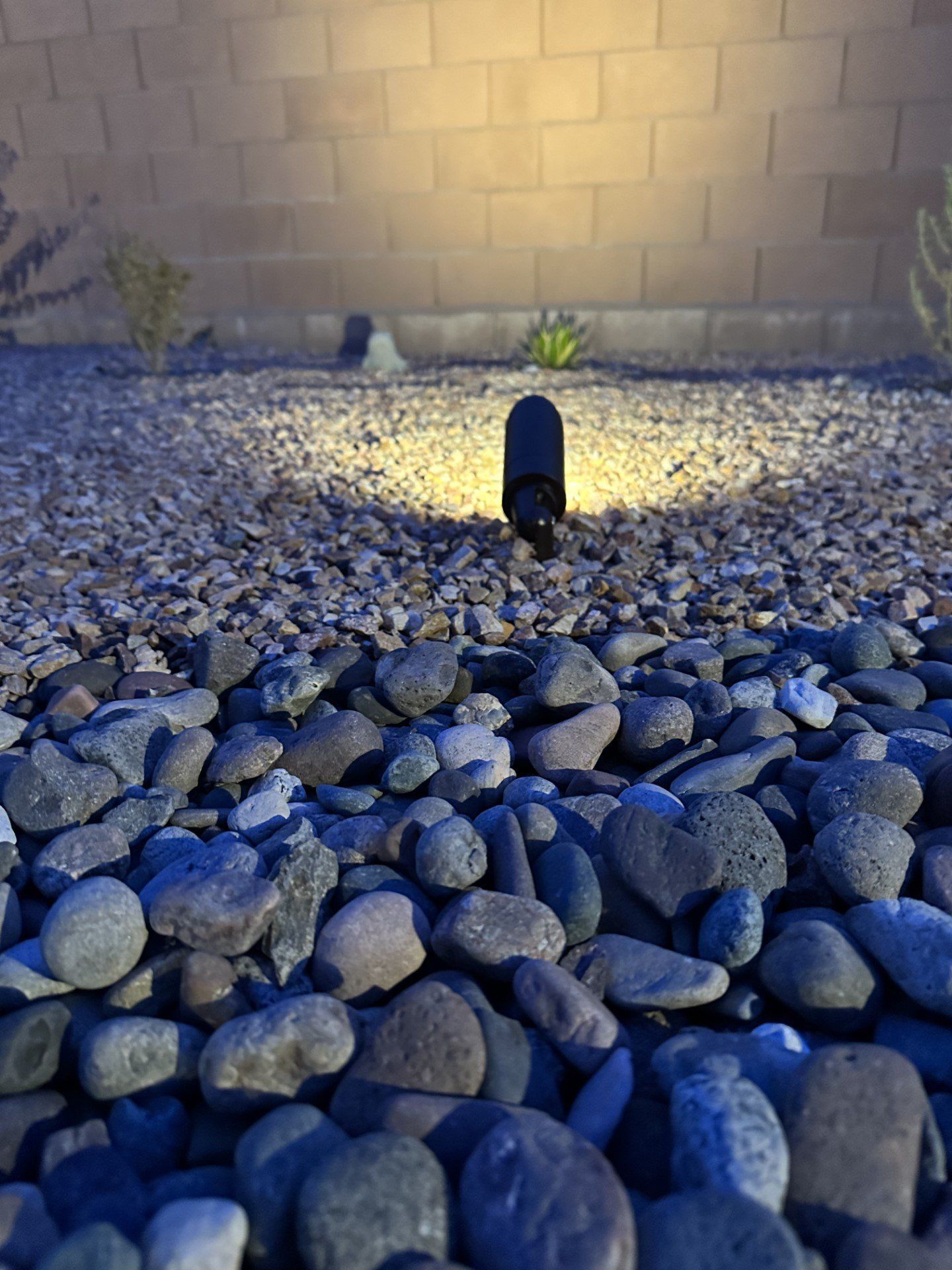 A black outdoor light illuminates rocks in a yard, with a tan wall in the background.