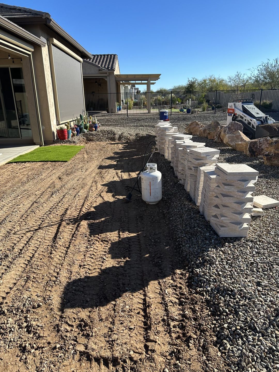 Backyard with building materials, propane tank, and prepared soil, sunny day.