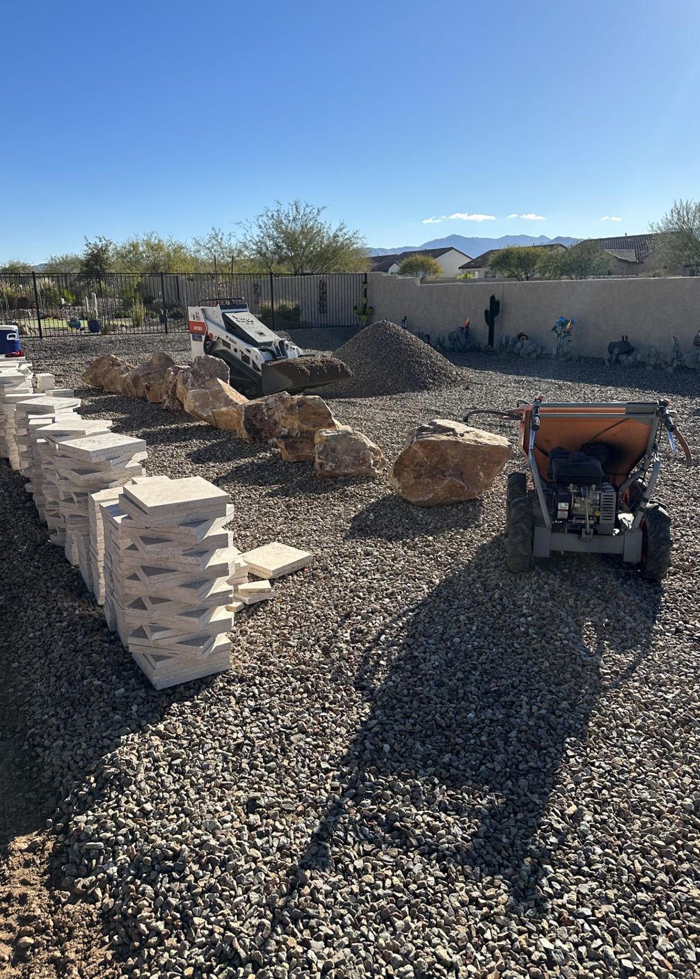 Construction site with stacks of pavers, rocks, gravel, and small machine under a blue sky.
