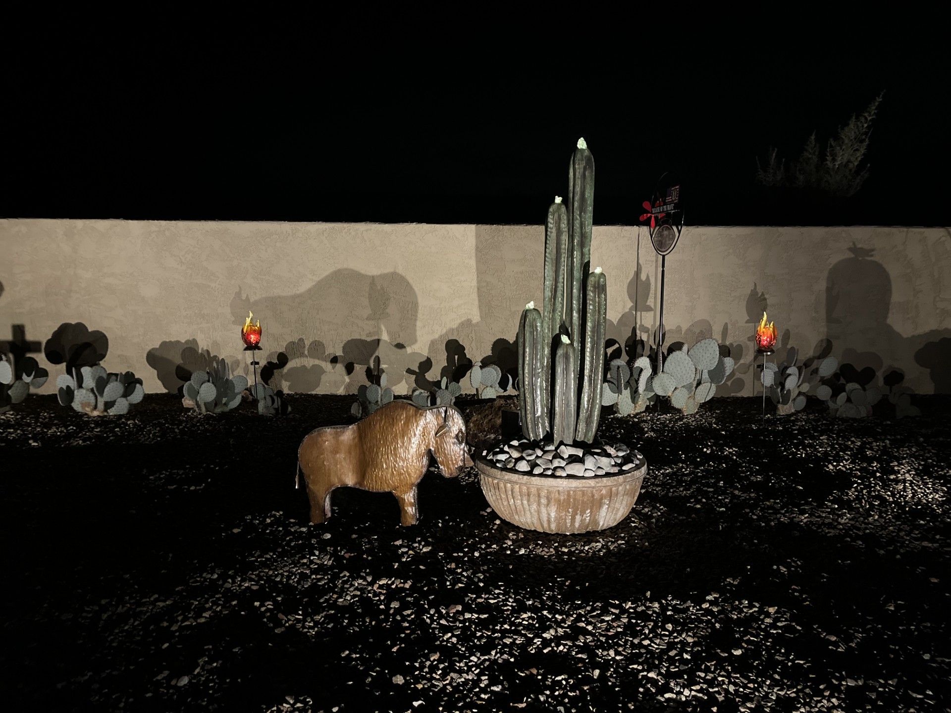 Statue of a pony in a dark yard, with cactus and wall with lights.