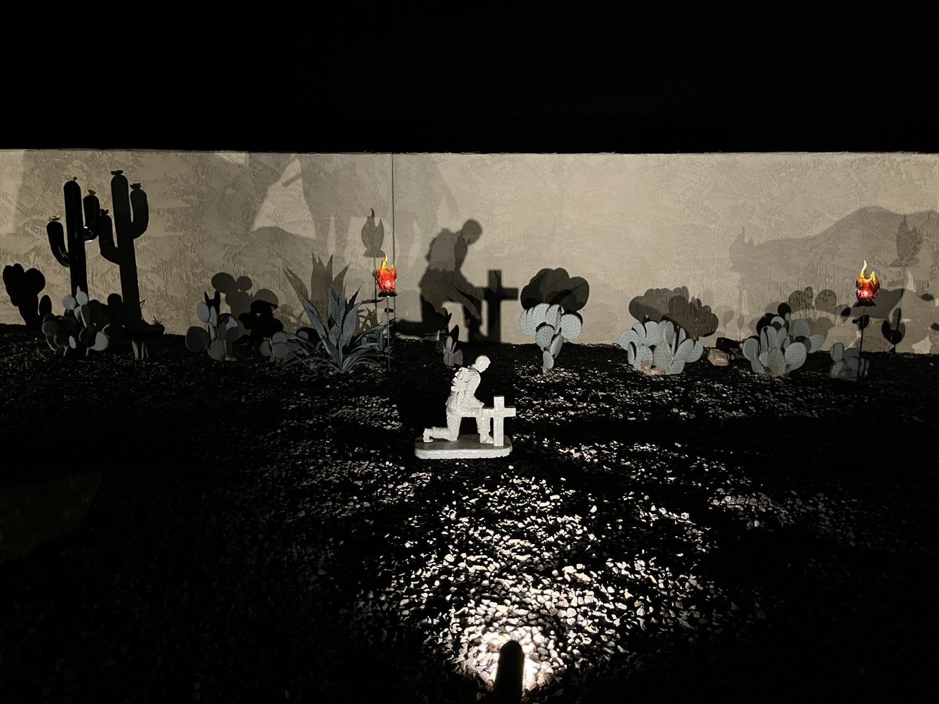 Desert scene: figure kneels by a cross. Another sits on a wooden structure; candles glow. Monochrome, dramatic lighting.