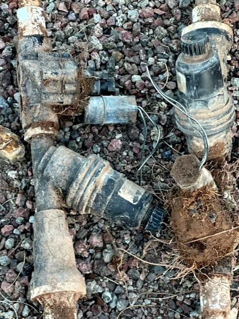 Irrigation system components on gravel: valves, filter, and fittings, covered in dirt.