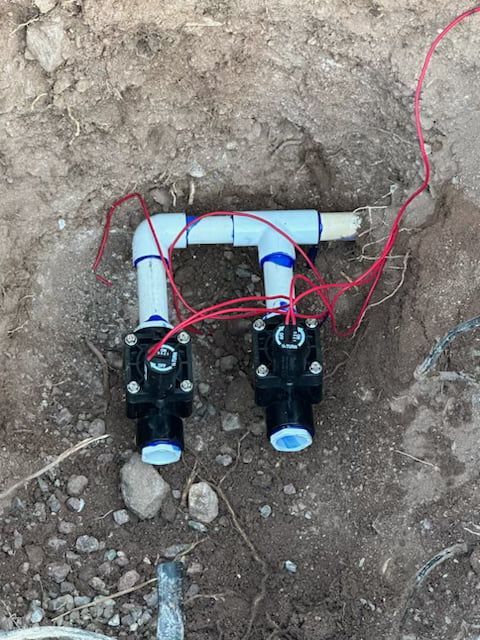 Two black sprinkler valves connected by white PVC pipes in a dirt trench, red wires attached.