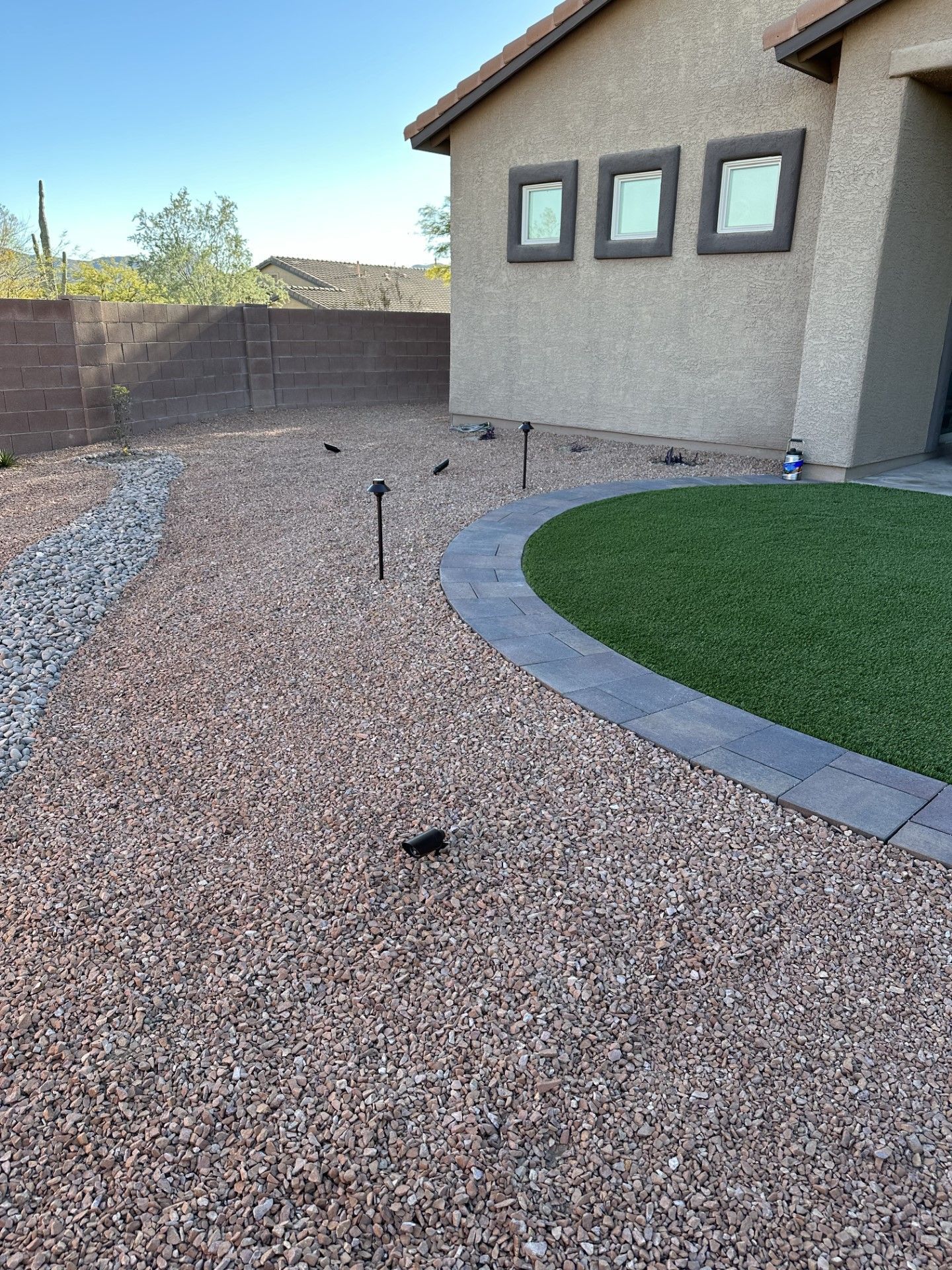 Gravel-covered side yard with small black lights, a patch of green grass, and a stucco house under a blue sky.
