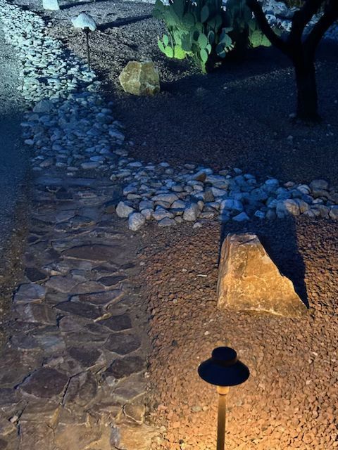 A lit stone pathway curves through a garden bed of rocks and mulch at night.