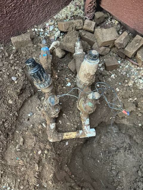 Irrigation system valves in a dirt hole near a building's wall and scattered bricks.