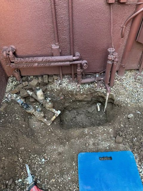 Hole dug in dirt near a building with exposed pipes, tools, and a blue kneepad. Brown tones.