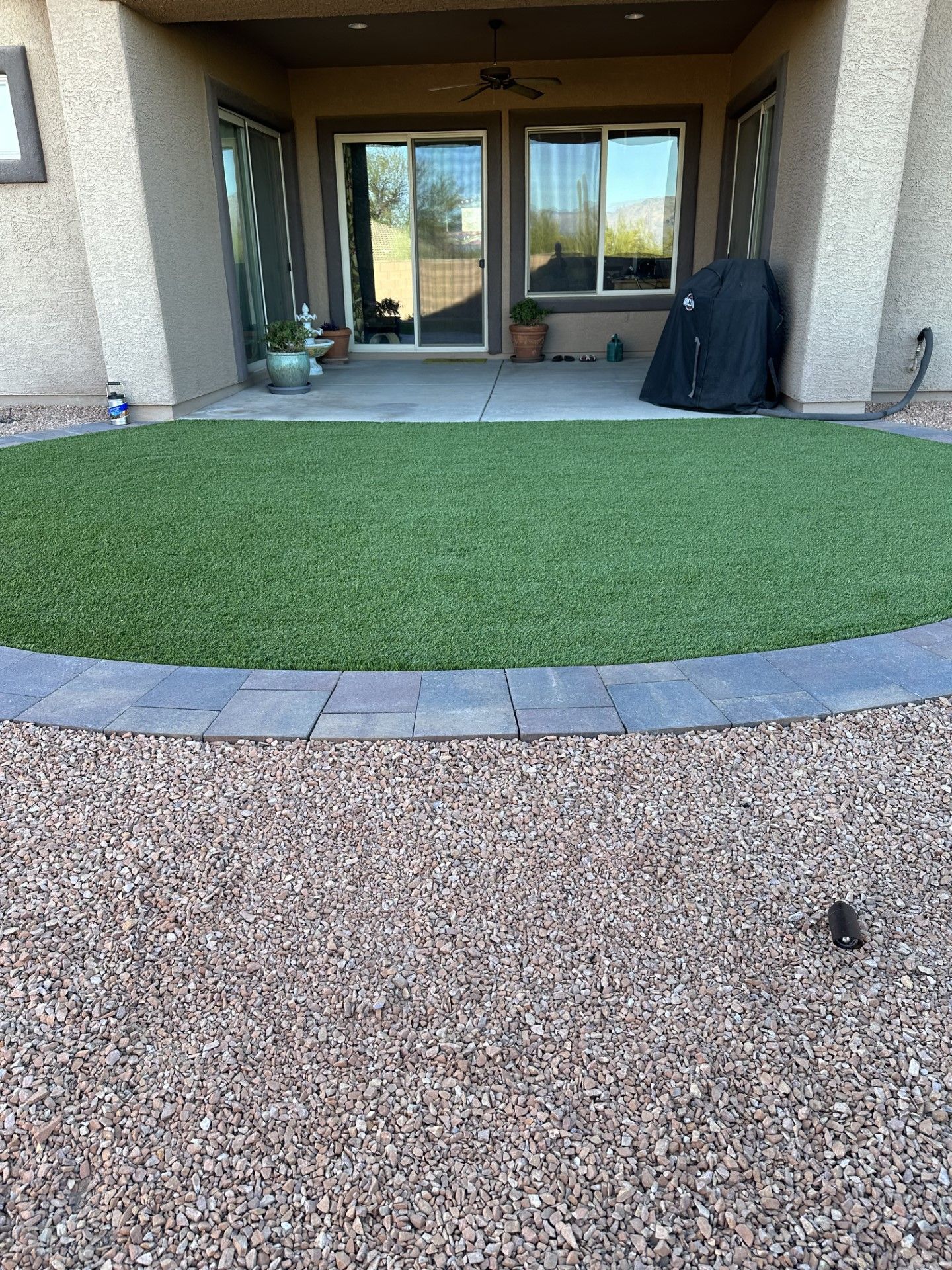 Backyard with artificial turf, bordered by paving stones and gravel. A covered patio is in the background.
