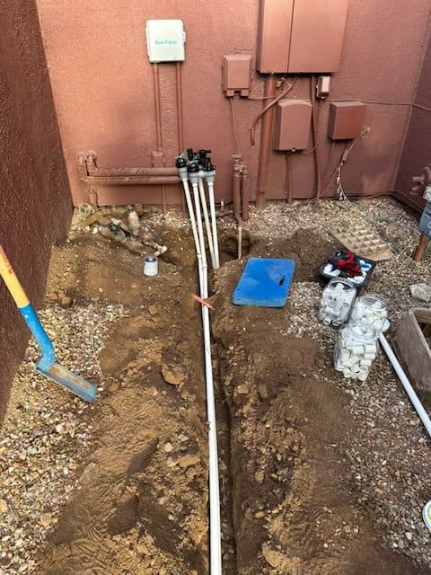 Trench with PVC pipes and sprinkler heads in front of a building with electrical boxes. Shovel and tools visible.