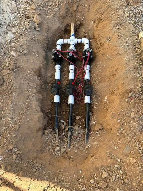 Irrigation valve manifold installed in a dirt trench, with three black valve assemblies connected to white PVC piping.