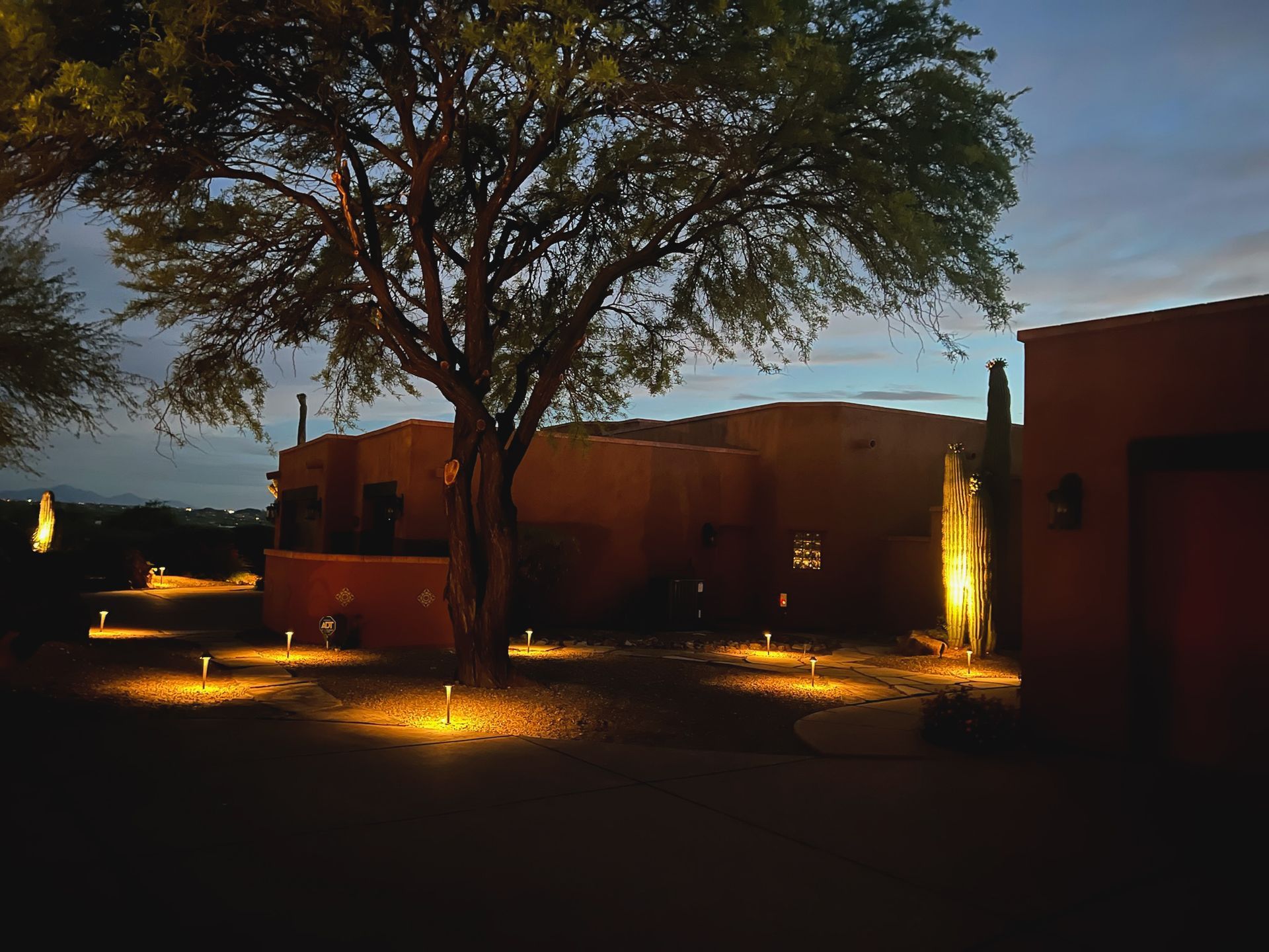 Exterior nighttime view of a southwestern-style home with landscape lighting illuminating the walkway and a large tree.