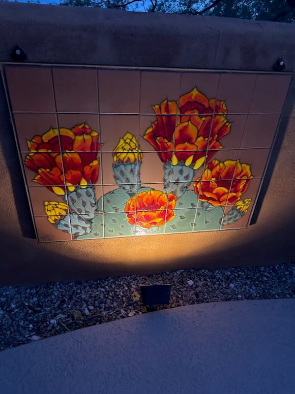 Tile mural of flowering cacti with orange blooms, mounted on a tan stucco wall, illuminated by a spotlight.