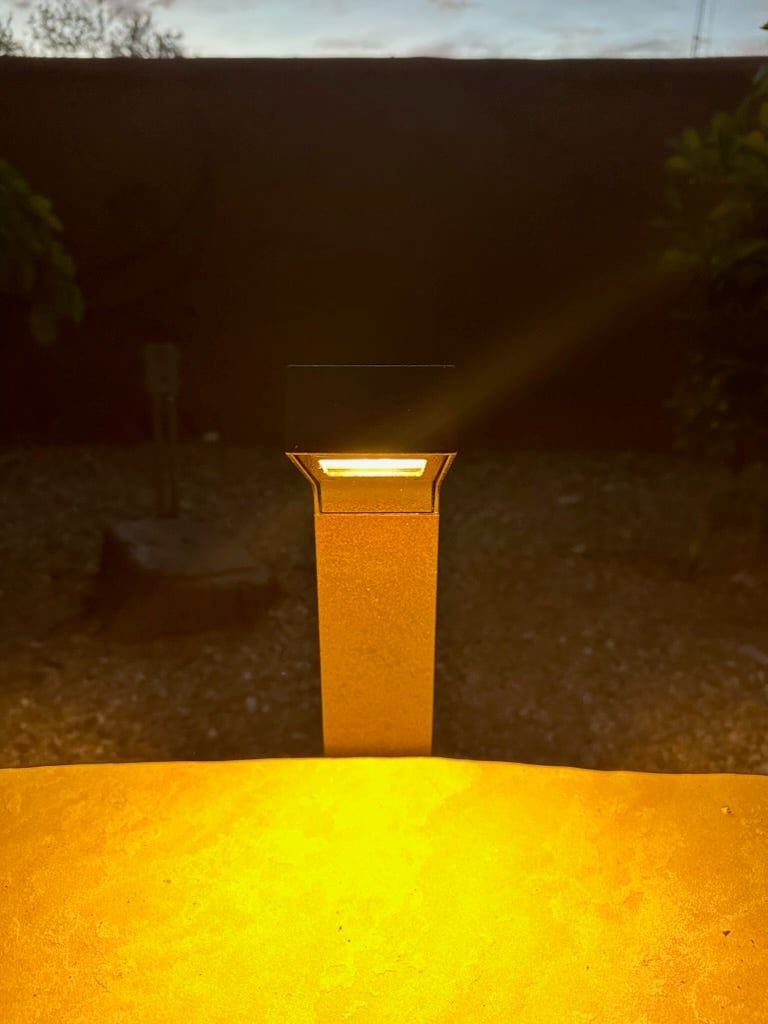 Lit, rectangular outdoor lamp casting warm yellow light on ground and wall.