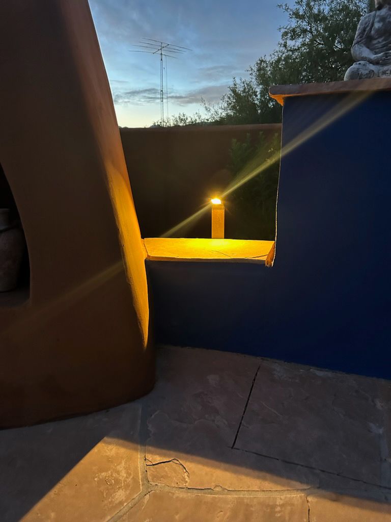 A lit outdoor light on a blue wall at dusk, beside an arched brown structure, and stone tiles.