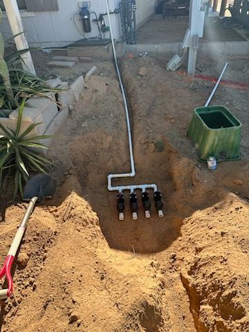 Dug trench with buried irrigation system; black valves, PVC pipes, green junction box, shovel nearby.