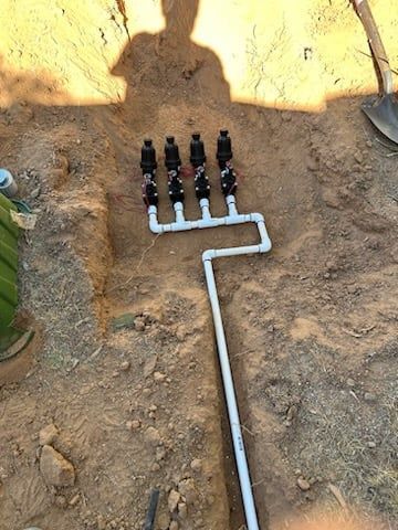 Irrigation system with 6 black valves connected to white PVC pipes in a dirt trench.