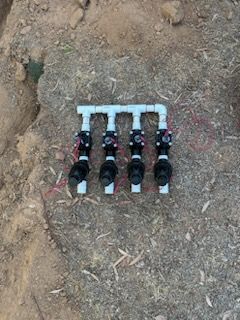 Four black sprinkler valves connected by white PVC pipes on dirt.