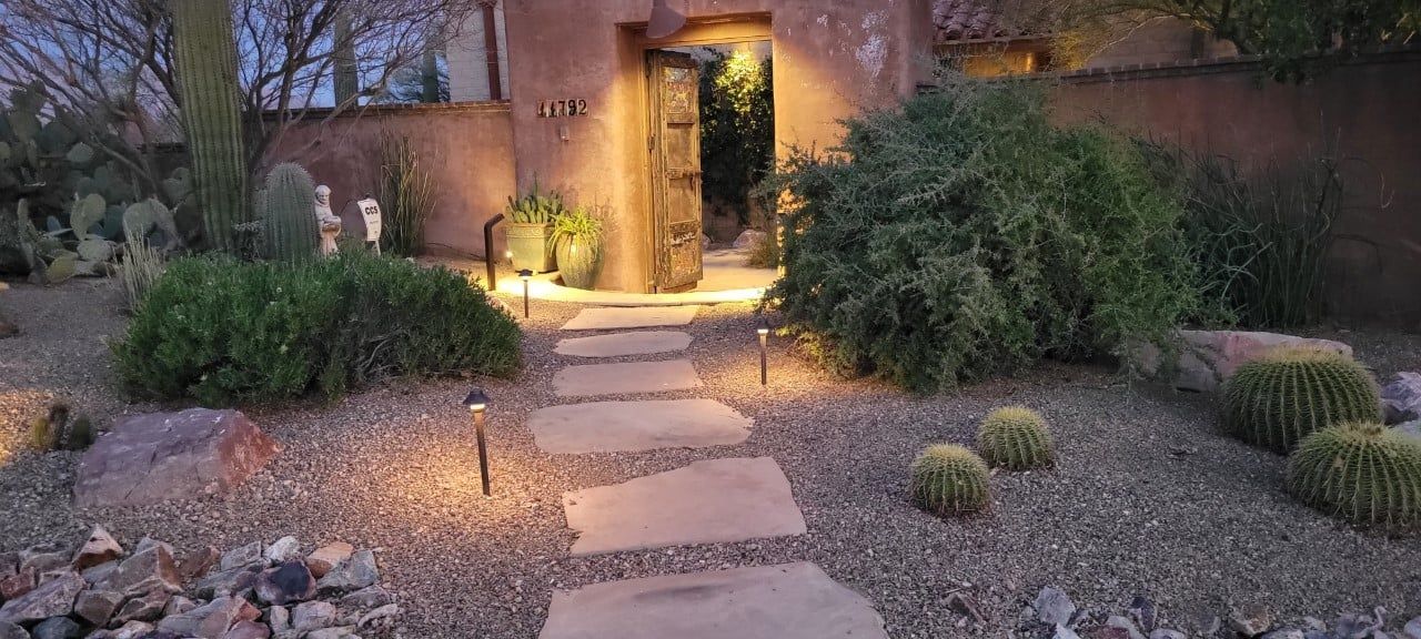 Stone path leads to a wooden door, illuminated by outdoor lights and surrounded by greenery and gravel.