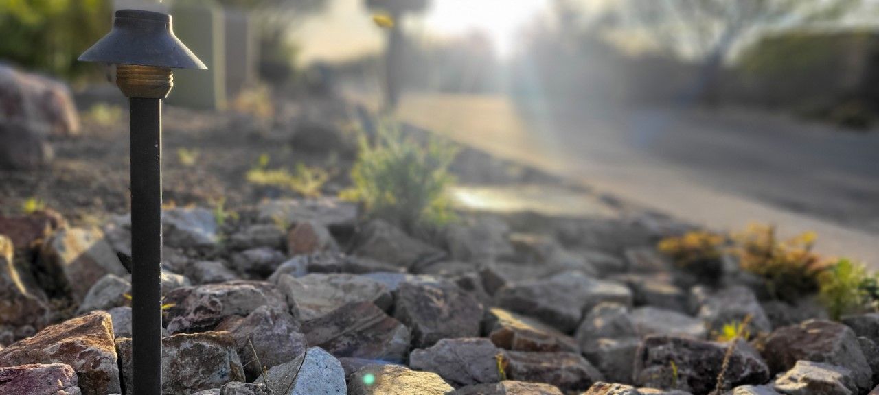 Black lamp post with a rocky landscape and a blurred road in the background, sun glare.