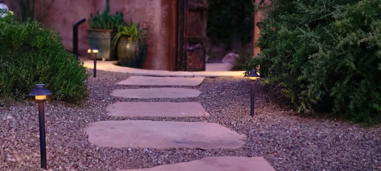 Stone path illuminated by low lights leads to a wooden door and lush greenery.
