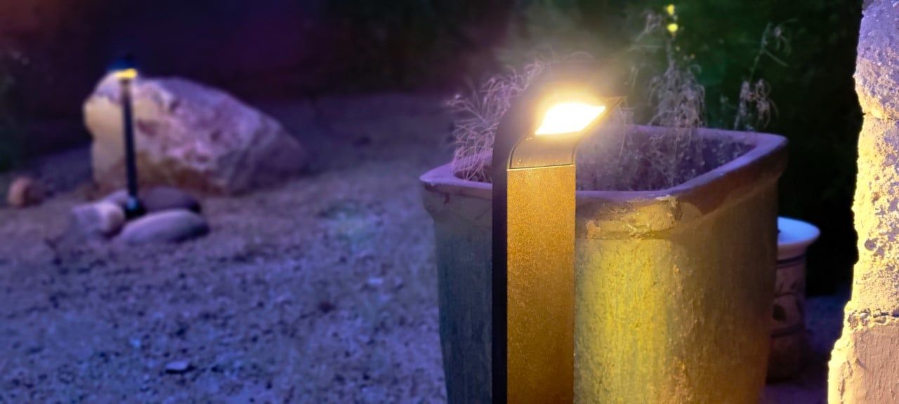 Outdoor scene with lit lights illuminating a large stone, a plant pot, and a stone wall at night.