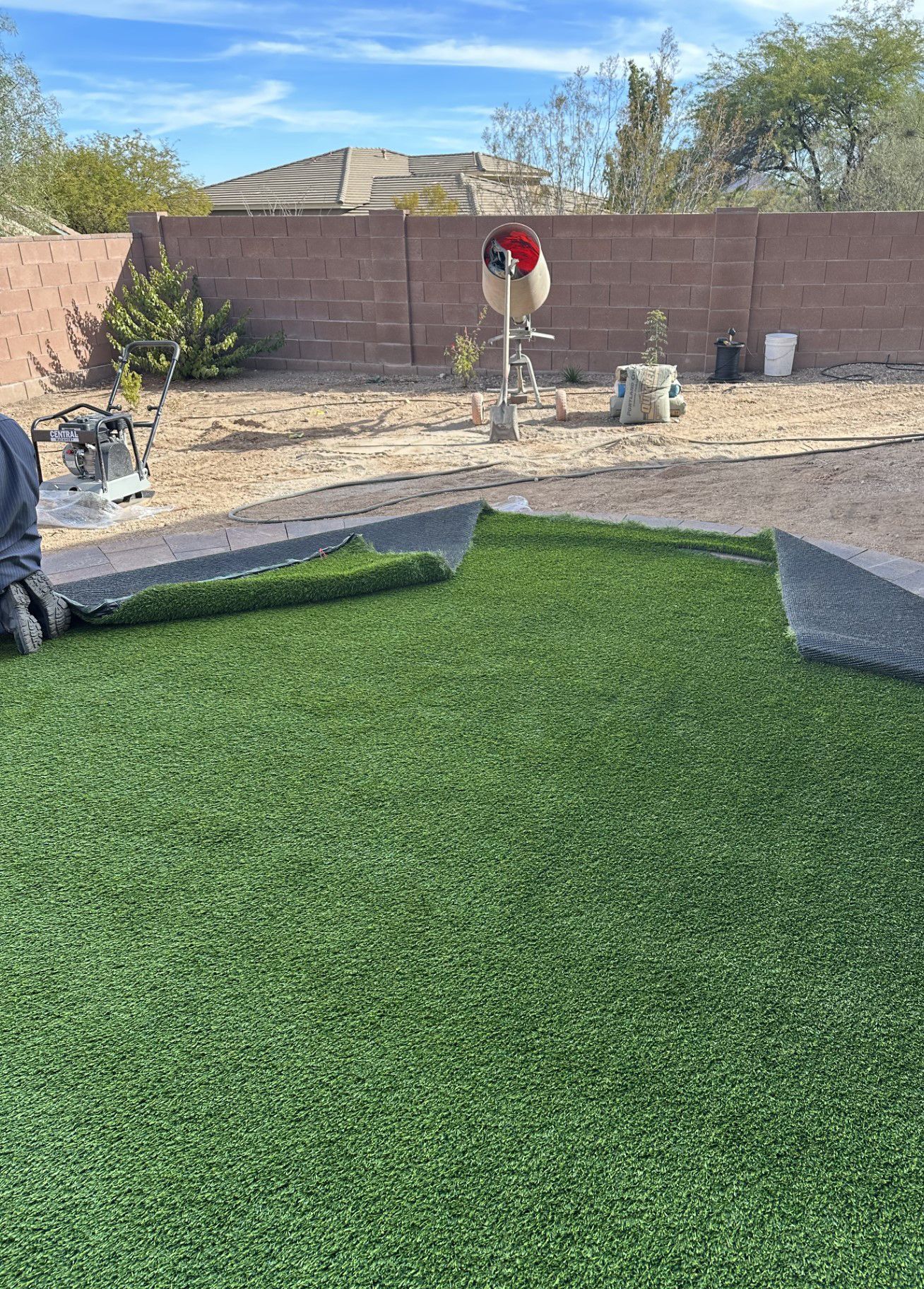 Laying artificial turf in a backyard. Rolls of green turf are unrolled on dirt, in front of a brick wall and vegetation.