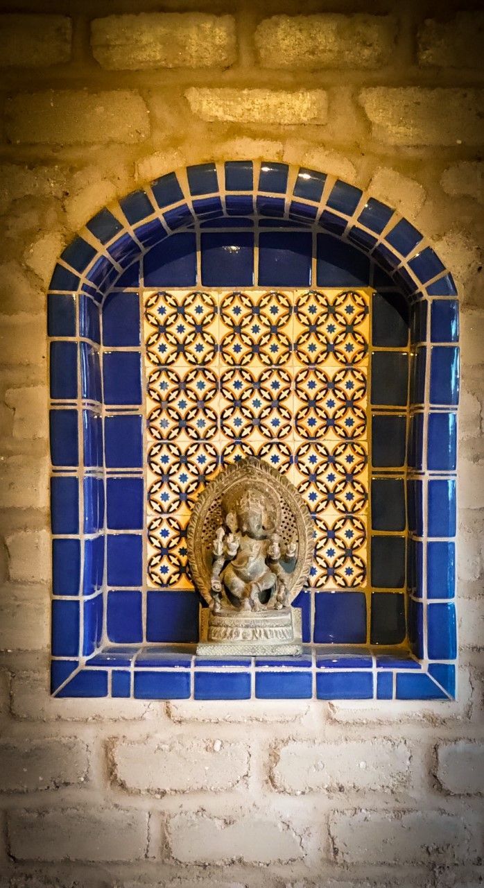 Stone statue of deity in a blue-tiled alcove with patterned tile background, set in a brick wall.